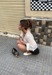 Valkoinen, puoliksi läpinäkyvä paita, jossa on kellotakasulet, mustapilkullinen hame, valkoiset tennarit ja kuvioitu käsilaukku teksturoidulla päällysteellä.