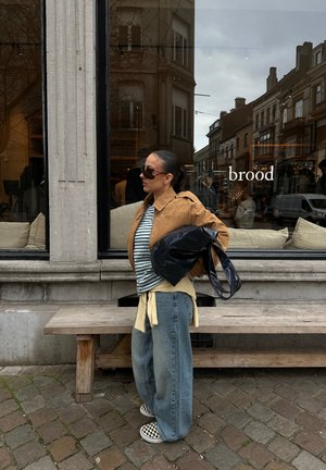 Mujer con gafas de sol, chaqueta marrón, camiseta de rayas y vaqueros anchos, llevando un bolso grande negro, parada frente a una cafetería con la palabra "brood" en la ventana.