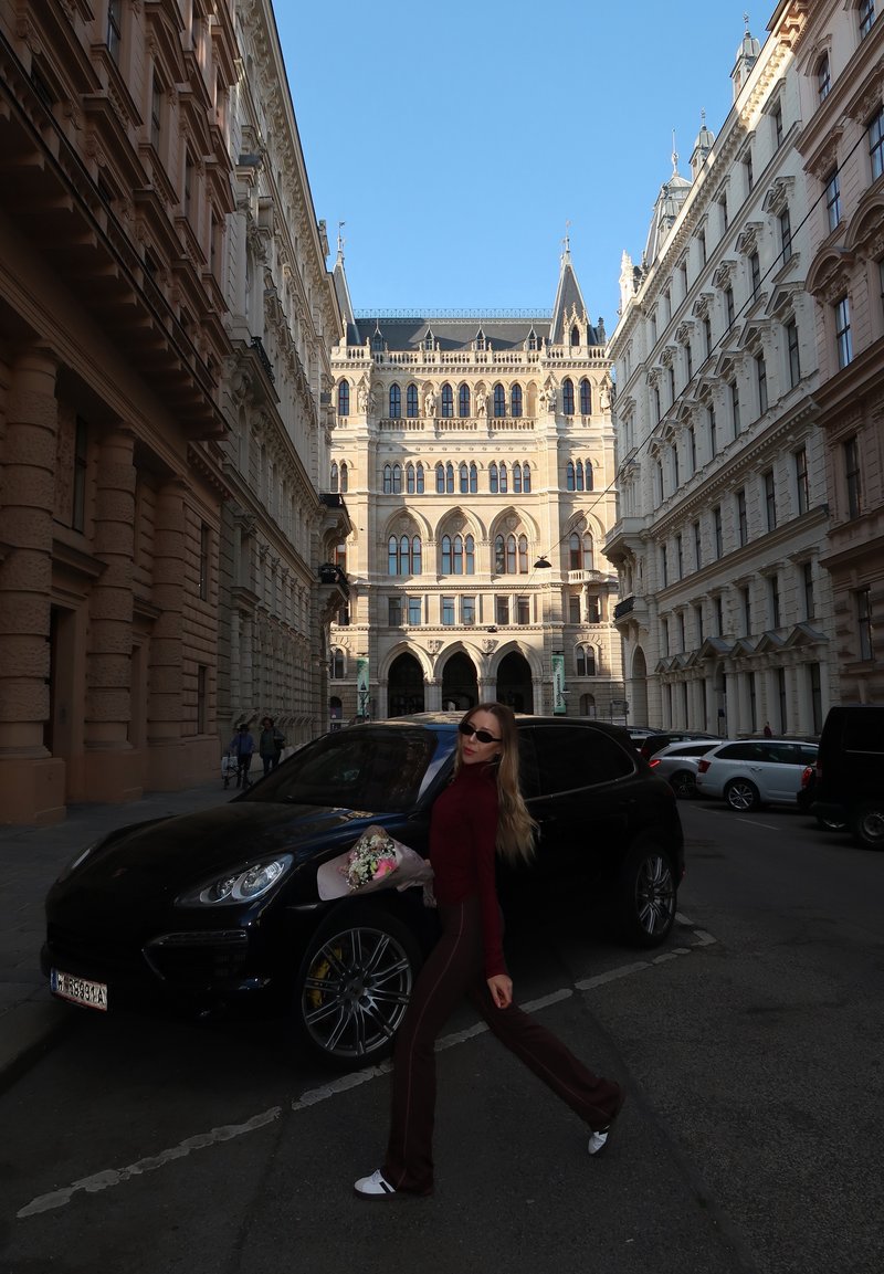 Frau mit Sonnenbrille geht an einem schwarzen Auto auf einer von historischen, ornamentierten Gebäuden gesäumten Stadtstrasse unter klarem blauem Himmel vorbei.