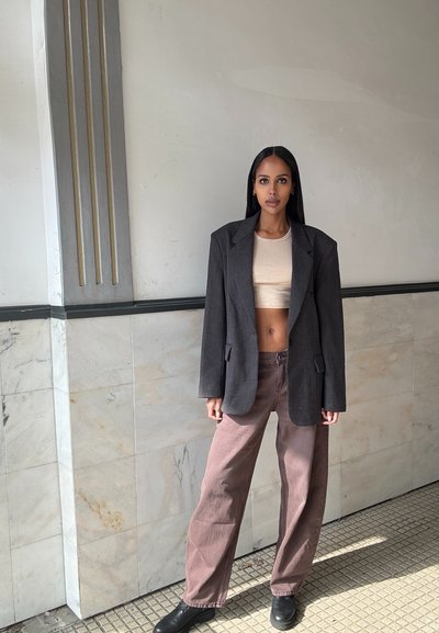 Blazer gris de gran tamaño sobre un crop top beige, combinado con pantalones sueltos marrones y zapatos negros. El entorno tiene paredes de mármol y acentos geométricos.