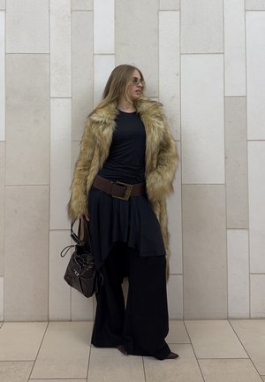 Abrigo de piel sintética en marrón claro, blusa negra, falda negra fluida, pantalones anchos y cinturón marrón con hebilla metálica. Sostiene un bolso de tela oscura.