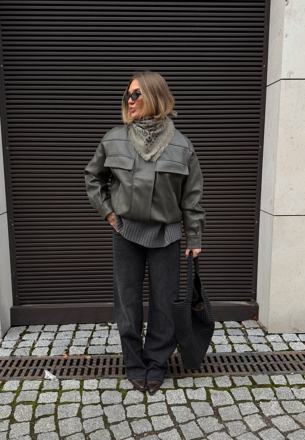 Veste en cuir verte avec grandes poches frontales, pull gris texturé, pantalon large sombre, et sac tressé noir.