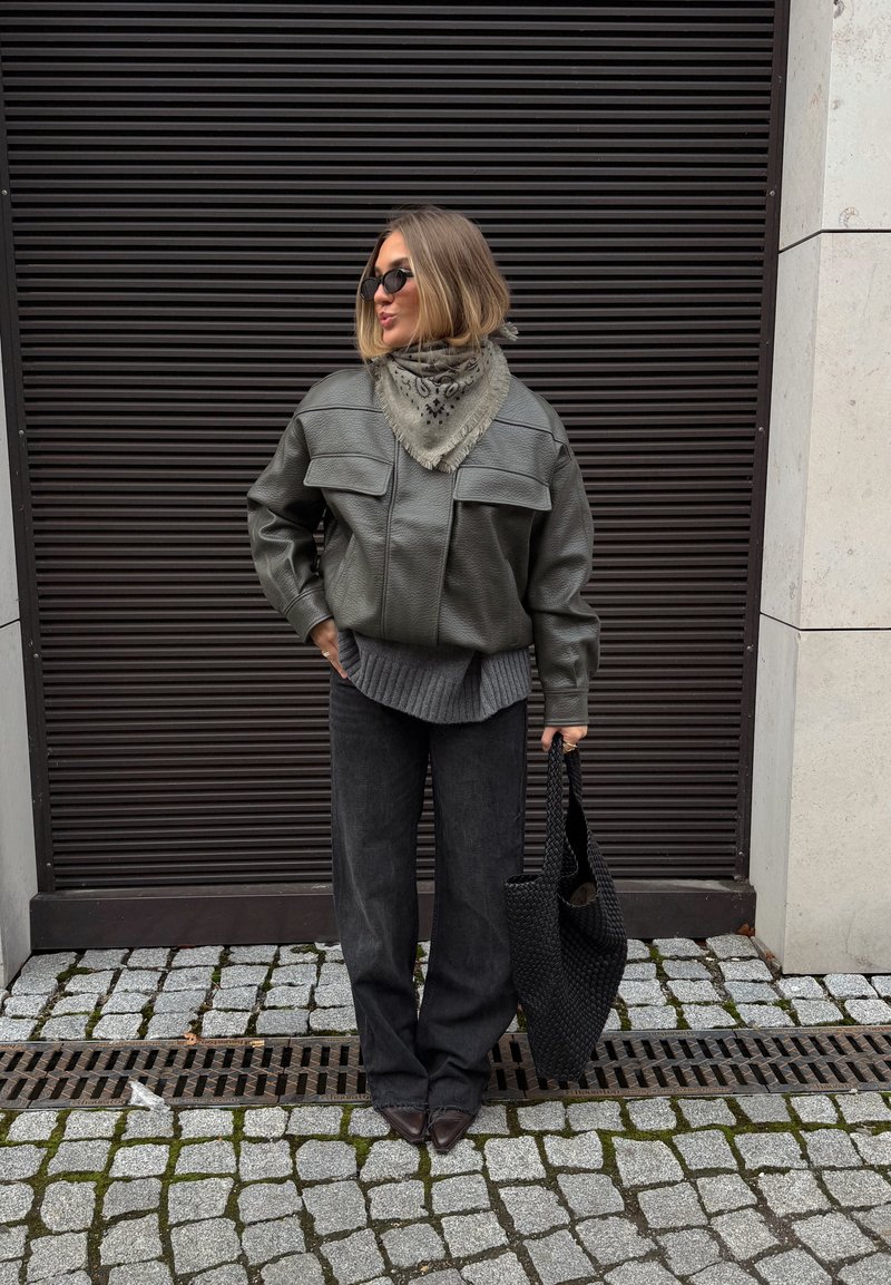 Veste en cuir verte avec grandes poches frontales, pull gris texturé, pantalon large sombre, et sac tressé noir.