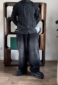 Personne portant une chemise rayée noire et blanche, un jean large noir et des chaussures noires à plateforme épaisses, debout à l'intérieur sur un sol en bois.