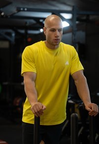 Svetlo rumena športna majica s kratkimi rokavi z logotipom Adidas, kombinirana s črnimi kratkimi hlačami. Oseba je postavljena ob vadbeno opremo.