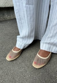 Πόδια που φορούν διαφανείς μπεζ πλέκτες μπαλαρίνες, με βαμμένα ν�ύχια ποδιών, συνδυασμένα με λευκά παντελόνια με ρίγες και φαρδιά κόψιμο, σε επιφάνεια άσφαλτου.