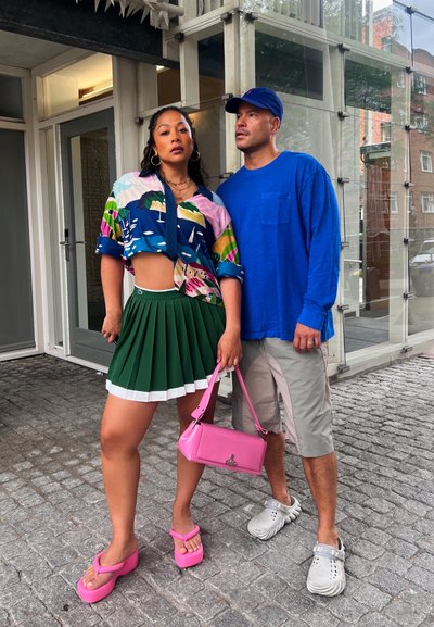 Camisa corta colorida con un fondo azul y patrones multicolores, falda plisada verde, bolso rosa y sandalias de plataforma. Camisa de manga larga azul para hombres y pantalones cortos claros.
