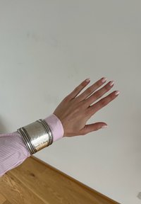 Srebrna manžetna narukvica s kovanom teksturom, nosi se preko ružičaste i bijele prugaste košulje. Ima glatku površinu i više kružnih naglasaka.
