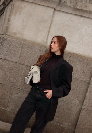 Woman with long red hair wearing black blazer and pants, holding light gray handbag, standing against stone wall, looking sideways.