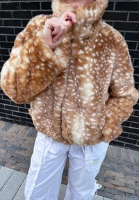 Casaco de pele sintética em castanho claro com padrão de manchas brancas, gola alta, fecho éclair frontal e textura felpuda. Usado com calças brancas.