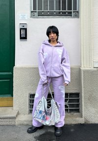 Personne portant un sweat à capuche lavande et un pantalon assorti avec des motifs blancs, tenant un grand sac à main gris texturé, debout près d'un mur crème.