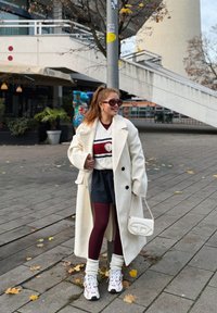 Palton de lână albă, pulover cu dungi și accente bordo, pantaloni scurți cu dungi fine, colanți bordo, geantă albă și adidași groși cu detalii colorate.