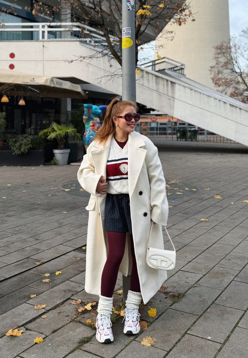 Manteau en laine blanche, pull rayé avec des accents bordeaux, shorts à fines rayures, leggings bordeaux, sac blanc et baskets épaisses aux détails colorés.
