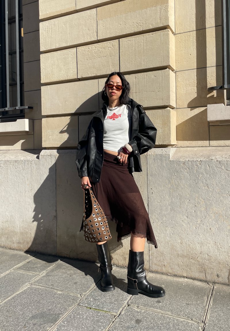 Femeie purtând ochelari de soare, tricou alb scurt cu imprimeu grafic, jachetă neagră din piele, fustă maro, cizme negre, ținând o geantă maro cu ochiuri metalice, sprijinindu-se de perete.