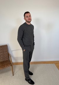 Homme portant un pull et un pantalon gris foncé, debout les mains dans les poches sur un tapis beige près d'une chaise tissée contre un mur blanc uni.