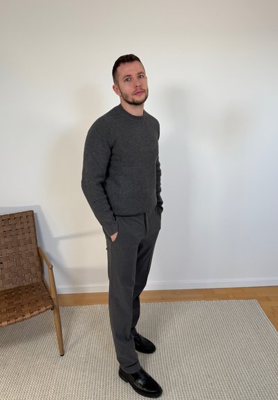 Hombre vestido con suéter y pantalones gris oscuro, de pie con las manos en los bolsillos sobre una alfombra beige cerca de una silla tejida contra una pared blanca lisa.