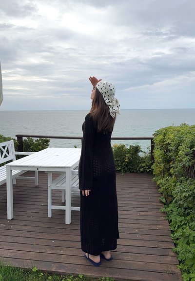 Vestido maxi negro de punto con mangas largas, llevado con un pañuelo blanco con lunares, de pie junto a una terraza de madera que da al mar.
