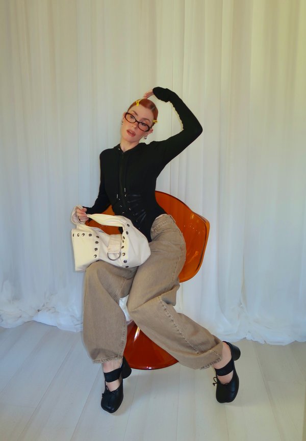 Una persona con gafas, un top negro y unos pantalones beige holgados posa en una silla naranja y sostiene un bolso blanco.