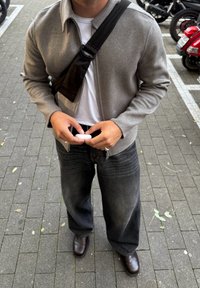 Férfi szürke dzsekit visel, fekete keresztpántos táskát hord, laza fekete farmernadrágot és barna cipőt, miközben egy kis fehér tokot tart egy járdán.