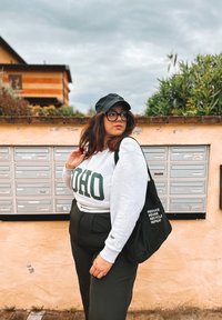 Sweatshirt cinza com texto verde "IDAHO", boné preto e calças pretas. Bolsa tote preta com texto branco. O fundo apresenta caixas de correio e plantas.