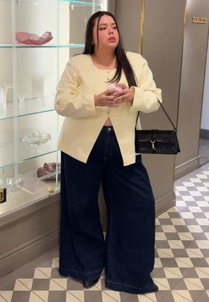 Mujer con blazer crema y pantalones anchos oscuros sostiene una taza, de pie cerca de estantes de vidrio con cuencos decorativos en un pasillo.