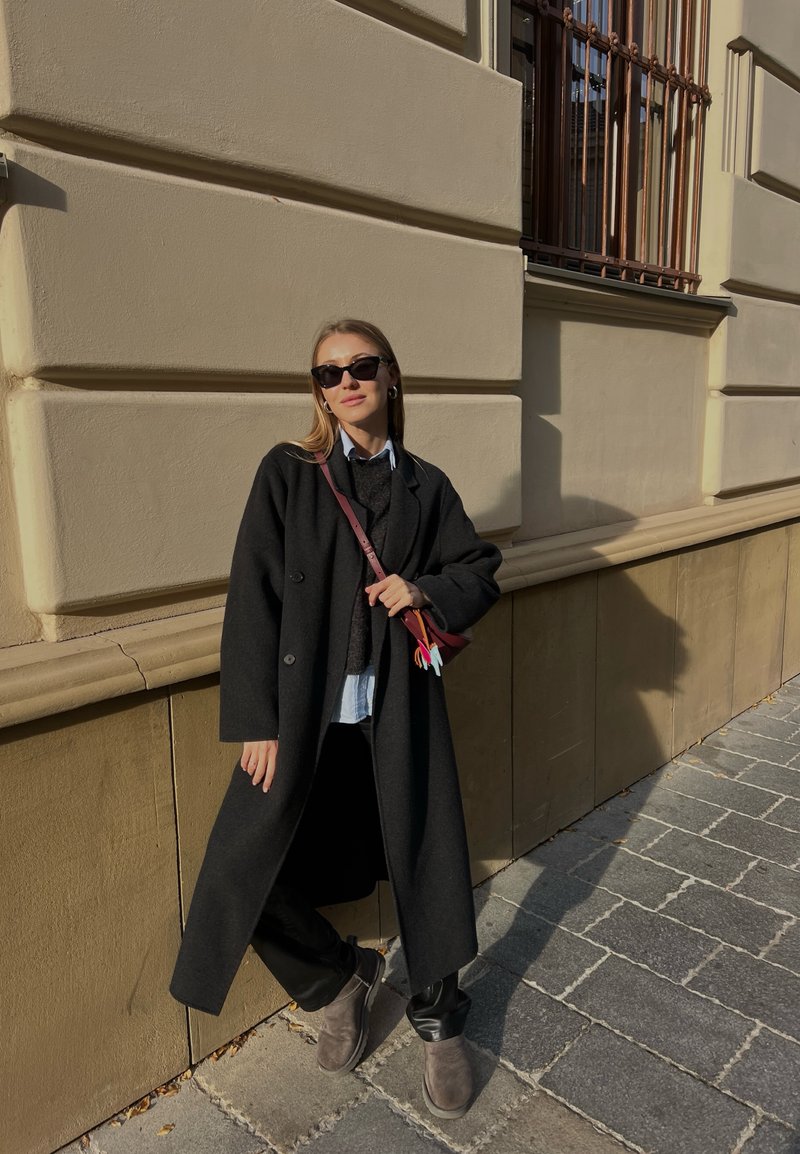 Μαύρο μάλλινο παλτό με μεγάλα κουμπιά, επάνω από γκρι πουλόβερ και ανοιχτό μπλε πουκάμισο. Μαύρο παντελόνι και γκρι μποτάκια μέχρι τον αστράγαλο; κρατάει μια μικρή τσάντα.