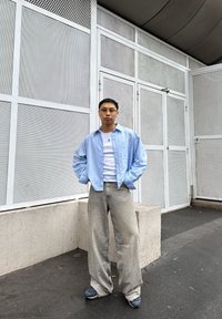 Világoskék vékony csíkos ing fehér póló felett, bőszabású szürke farmerrel és tengerészkék tornacipővel párosítva. Beton háttér és hálós panelek.