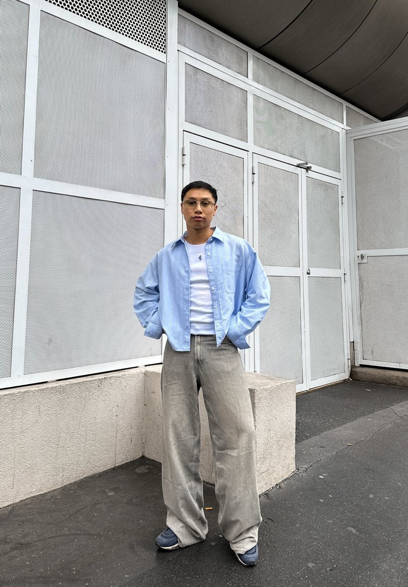 Camisa azul claro com riscas finas, abotoada, usada por cima de uma t-shirt branca, combinada com jeans cinzentos largos e ténis azul-marinho. Fundo de betão e painéis em rede.