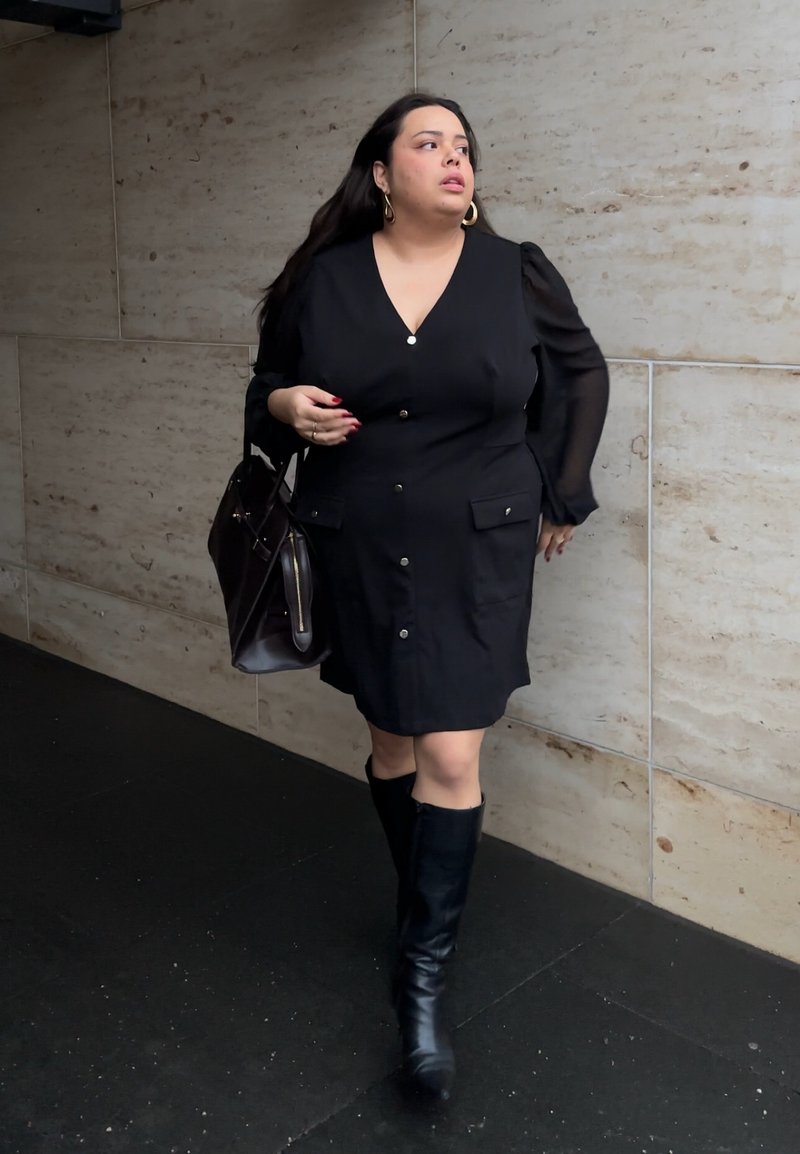 Robe noire boutonnée avec manches bouffantes, bottes noires jusqu'au genou et sac à main sombre. Tissu lisse et poches discrètes sur la robe.