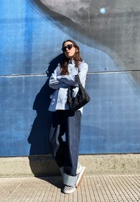 Camicia di jeans azzurro chiaro, borsa a spalla nera, pantaloni di jeans scuri larghi e sneakers slip-on a scacchi contro un muro blu.