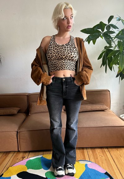 Top corto de tirantes con estampado de leopardo, chaqueta oversized marrón, jeans oscuros de campana, zapatillas blancas y negras, de pie sobre una alfombra circular multicolor.