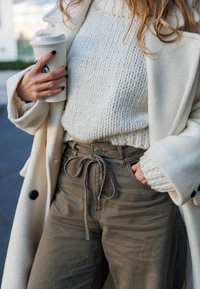 Abrigo de lana crema, suéter de punto blanco roto, pantalones marrones de cintura alta con cordón, sosteniendo un vaso de café desechable.