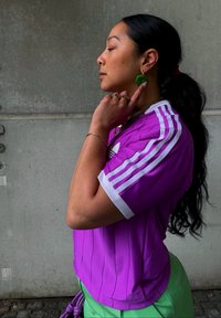 Maglia sportiva viola con strisce bianche, maniche corte e colletto a coste. Orecchini a cerchio verdi e anelli sovrapposti accentuano il modello.