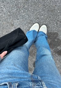 Personne portant un jean bleu clair et des chaussures blanches debout sur un bitume gris, tenant un sac noir dans la main gauche.