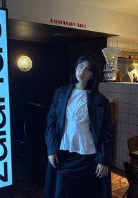 Manteau noir sur un haut péplum blanc, assorti  à une jupe noire. Cadre intérieur avec une lumière décorative et un mur carrelé visible.