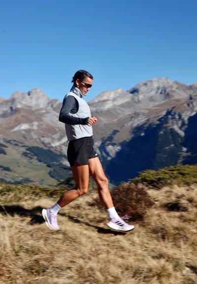 Mujer con gafas de sol y ropa deportiva corriendo cuesta abajo por un sendero montañoso con picos rocosos y un cielo despejado de fondo.