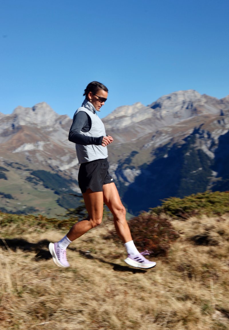 Vrouw in een zonnige bril en sportkleding die naar beneden rent op een bergpad met rotsachtige toppen en een heldere lucht op de achtergrond.