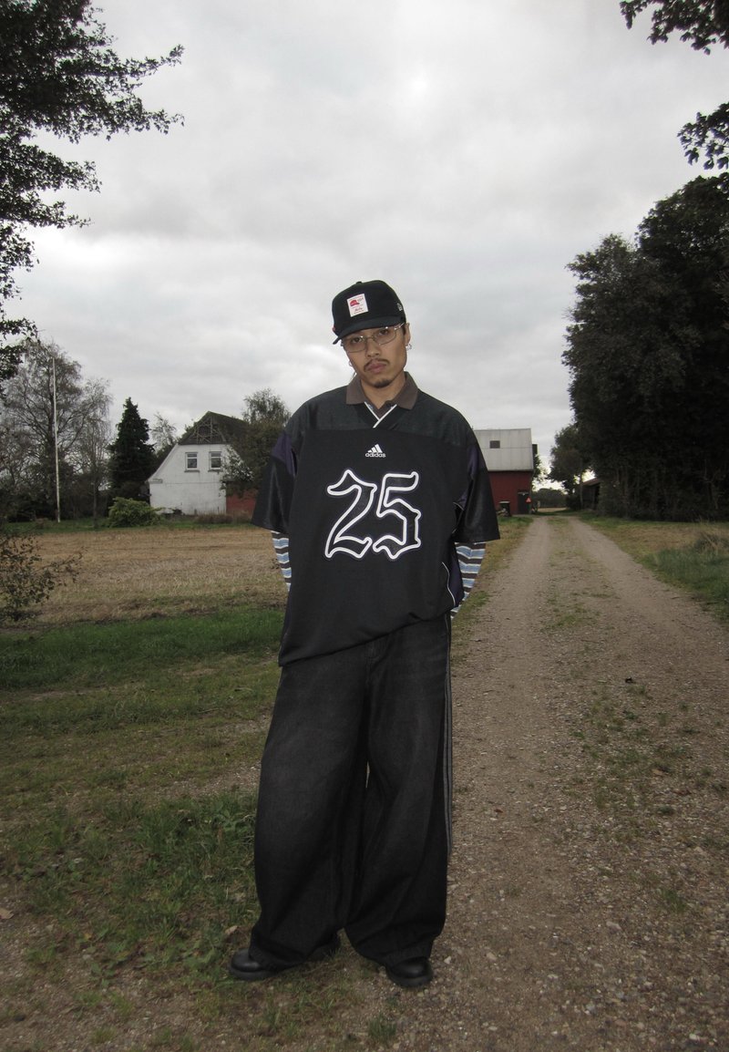 Black oversized jersey with white number "25," paired with wide dark jeans and a black cap. Striped long sleeves visible beneath the jersey.