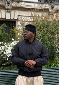 Čierna prešívaná bomber bunda s vysokým golierom a zapínaním na zips, kombinovaná s krémovými nohavicami. Doplnky zahŕňajú okrúhle okuliare a niekoľko prsteňov. V pozadí je zelené lístie a biele kvety.