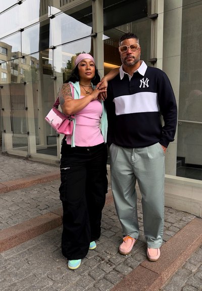 Dos personas posan al aire libre. Una lleva una camiseta rosa con una sudadera verde menta y pantalones cargo negros. La otra lleva un polo blanco y negro y pantalones verdes claros.