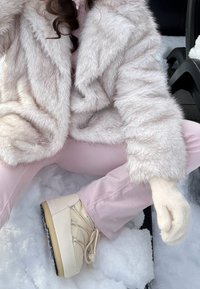 Jakna iz umetnega krzna kremne barve, svetlo rožnate oprijete hlače, bež zimski škornji z vezalkami, kremna pletena rokavica. V ozadju zasnežena podlaga.