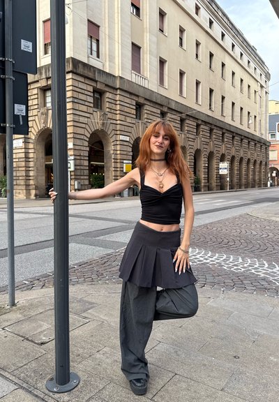 Top corto negro, falda plisada negra de capas y pantalones grises holgados. Los accesorios incluyen collares de capas y un gargantilla. Entorno urbano.