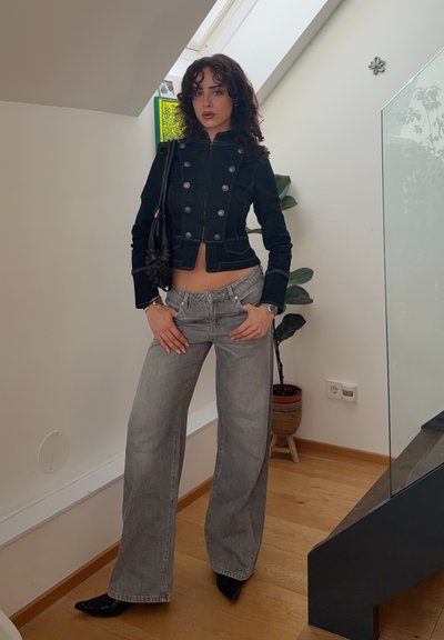 Joven mujer con chaqueta negra abotonada y vaqueros grises de pierna ancha, de pie en el interior cerca de una planta en maceta y una escalera.