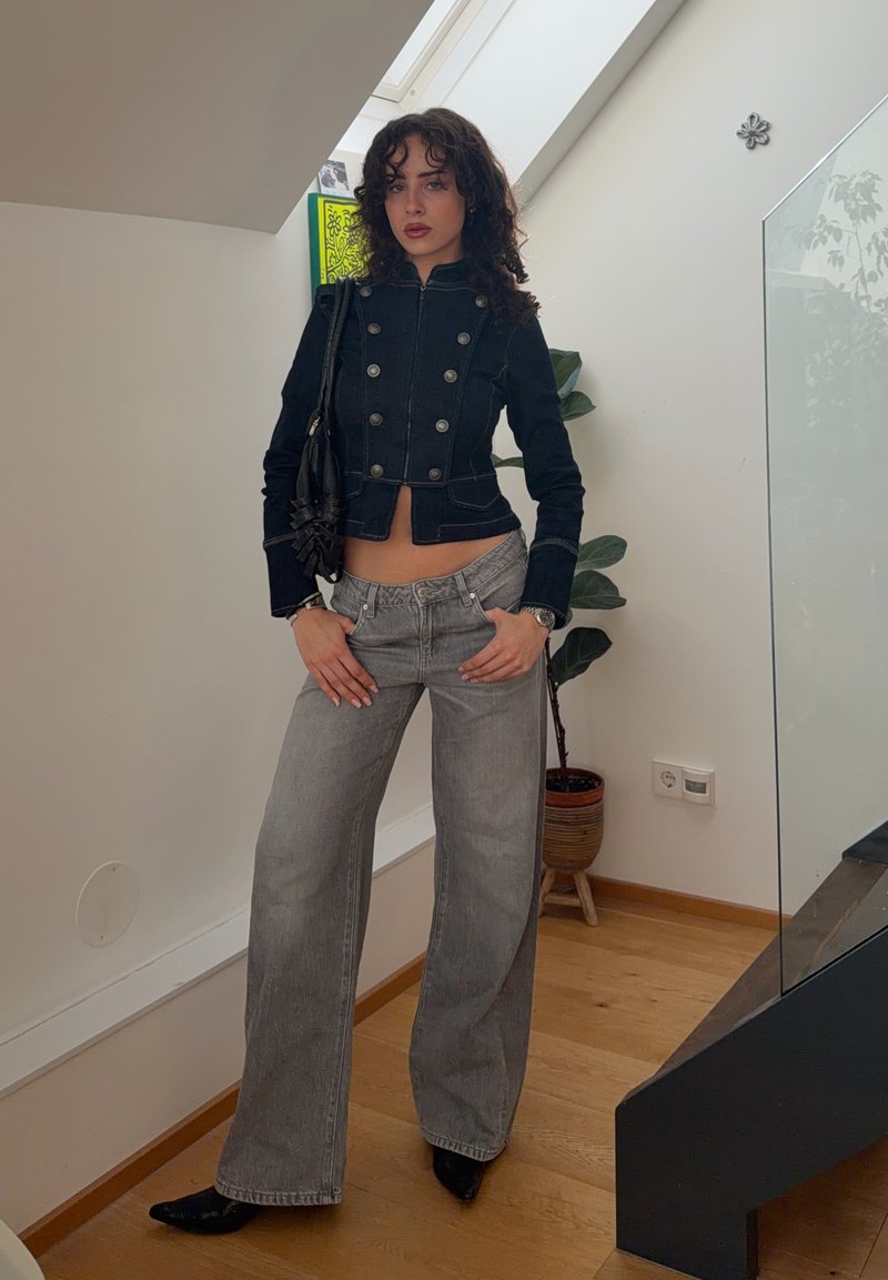 Mlada žena u crnoj jakni s gumbima i širokim sivim trapericama stoji u zatvorenom prostoru pokraj cvjetnog lonca i stepenica.