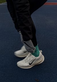 Fehér sportcipők fekete díszítésekkel, türkiz zoknival és fekete nadrággal párosítva. A háttér egy kék sportfelület.