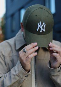 Person adjusting an olive green New York Yankees cap, wearing a beige suede jacket and a silver ring, in an urban outdoor setting.