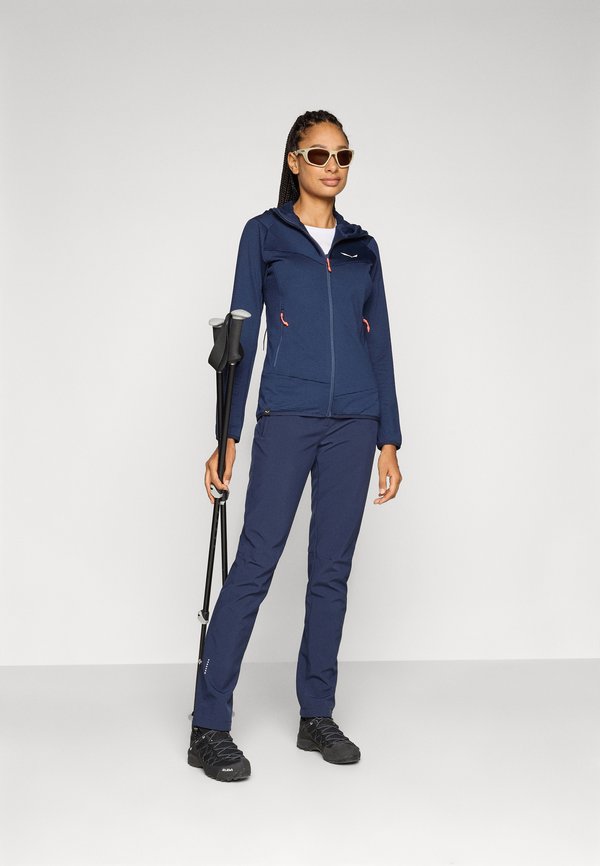 Marineblaue Zip-Jacke mit roten Akzenten, kombiniert mit passenden Hosen. Die Accessoires umfassen eine Sonnenbrille und Trekkingstöcke. Die Textur wirkt glatt.