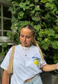 Witte graphic T-shirt met een citroenontwerp, gecombineerd met lagen gouden kettingen en een geweven beige tas, tegen een achtergrond van groene bladeren.