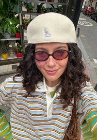 Biserna flis beret s logom kengura, uparena s plavim polo majicom sa zelenim i narančastim prugama. Vidljiva smeđa torba preko ramena.