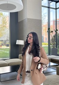 Γαλαζωπή καφέ σακάκι πάνω από λευκή μπλούζα, με στρωτή φούστα και μοτίβο τσάντας. Στρογγυλά λευκά γυαλιά ηλίου, μακριά σκούρα μαλλιά και ασημένια δαχτυλίδια. Σύγχρονος εσωτερικός χώρος.